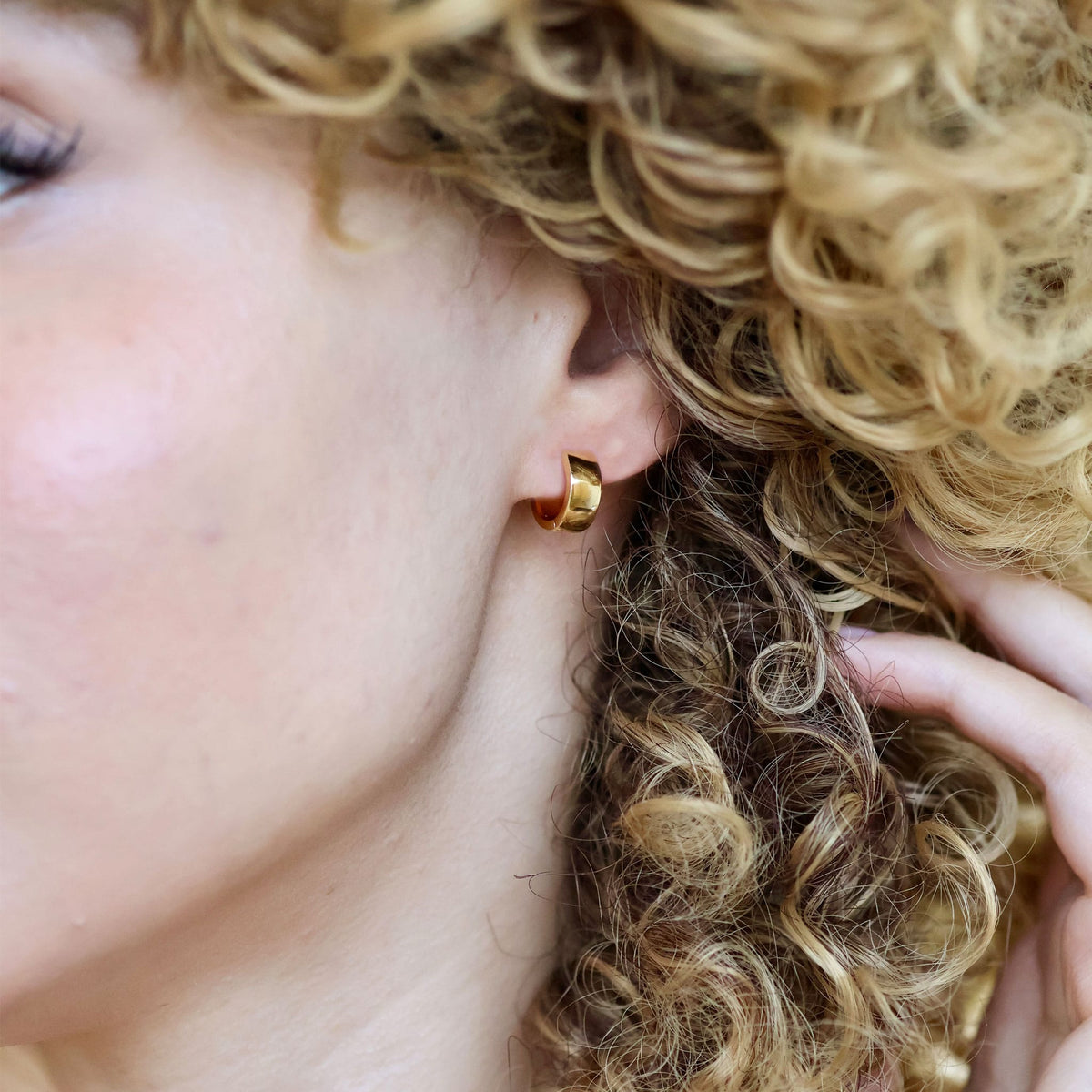 Woman with curly hair wearing a gold Wide Hinged Huggie Hoop