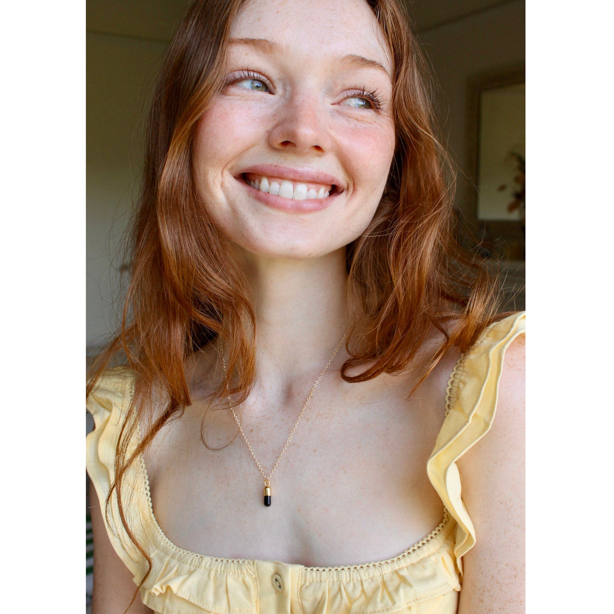 Smiling woman with long red hair wearing the Chill Pill necklace with a yellow top