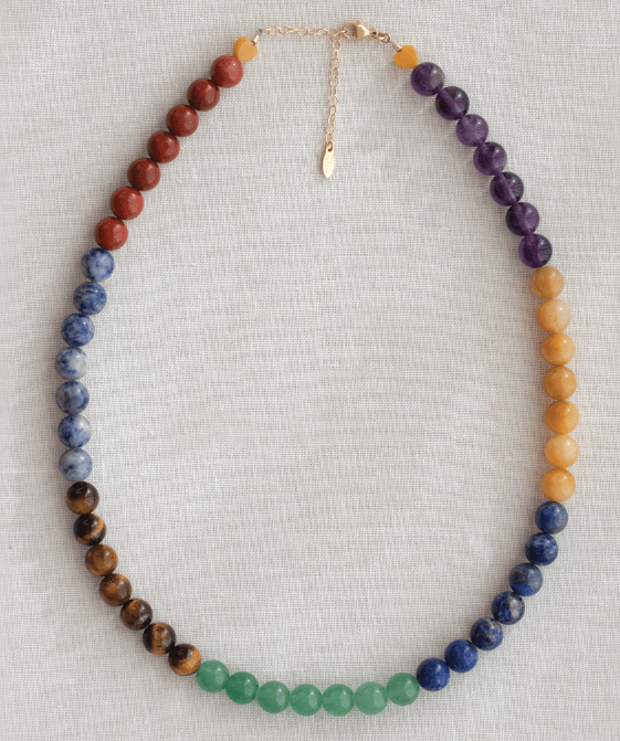 A beaded chakra necklace made of round gemstone beads in a rainbow sequence, laid flat on a light beige linen background. The necklace features orange, red, blue, brown, green, and purple beads, finished with a gold chain extender and clasp.