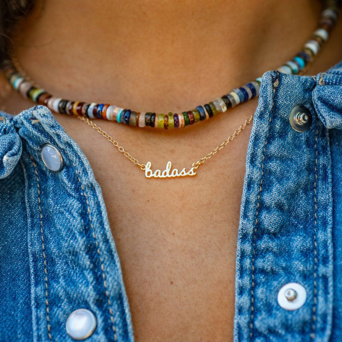 Gold Badass Charm Necklace layered with a multicolor beaded necklace and a denim jacket