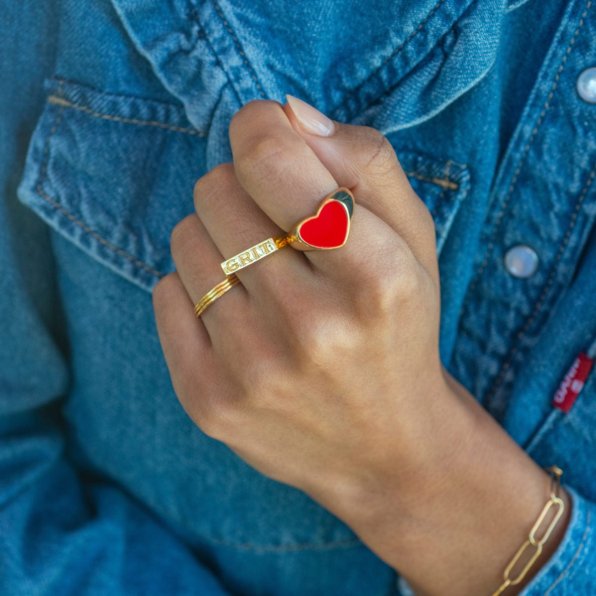 Hand on denim wearing the gold Grit Engraved Ring with a red heart ring