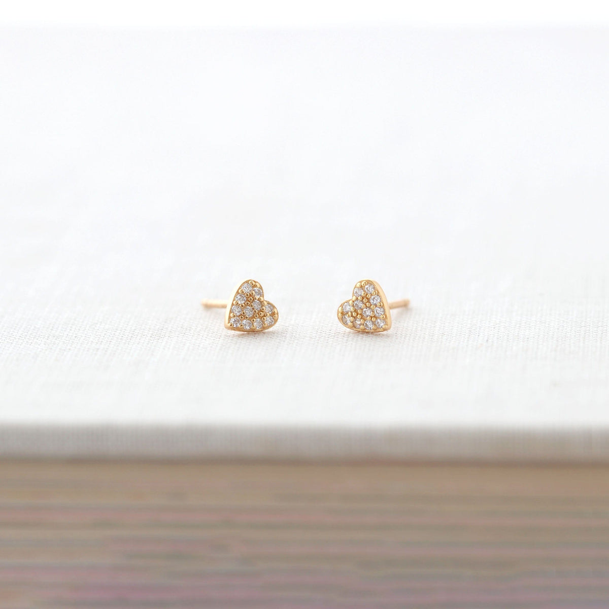 Gold heart crystal stud earrings resting on a white linen-covered book