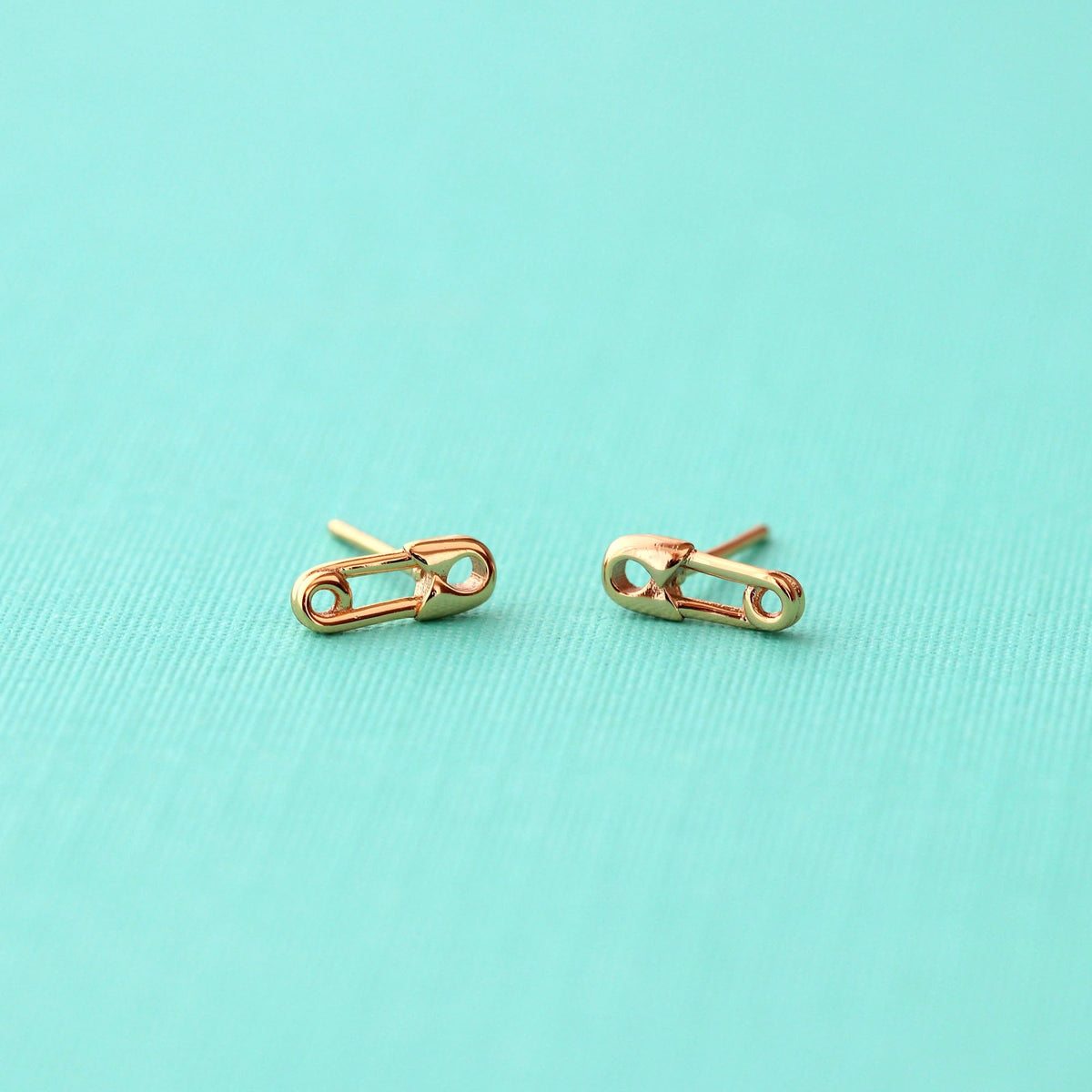 Gold Safety Pin Stud Earrings on an aqua background