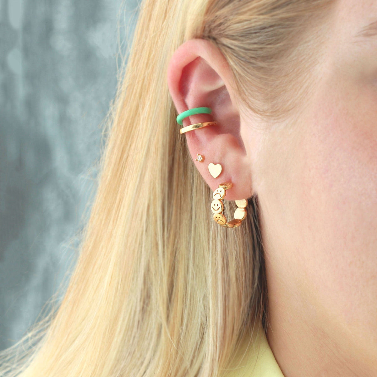 Hoop earrings styled on a model with blonde hair.  The hoops are in the first piercing, with a heart stud earring in second piercing and a cubic zirconia diamond stud in the third piercing with 2 ear cuffs on top- one is gold and the other is a turquoise color made from enamel.
