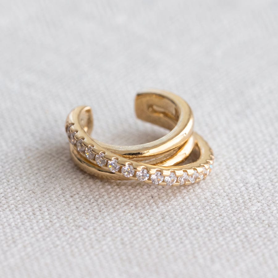 A gold huggie ring featuring a row of clear pavé-set round stones along the front band, photographed on a textured off-white fabric background.