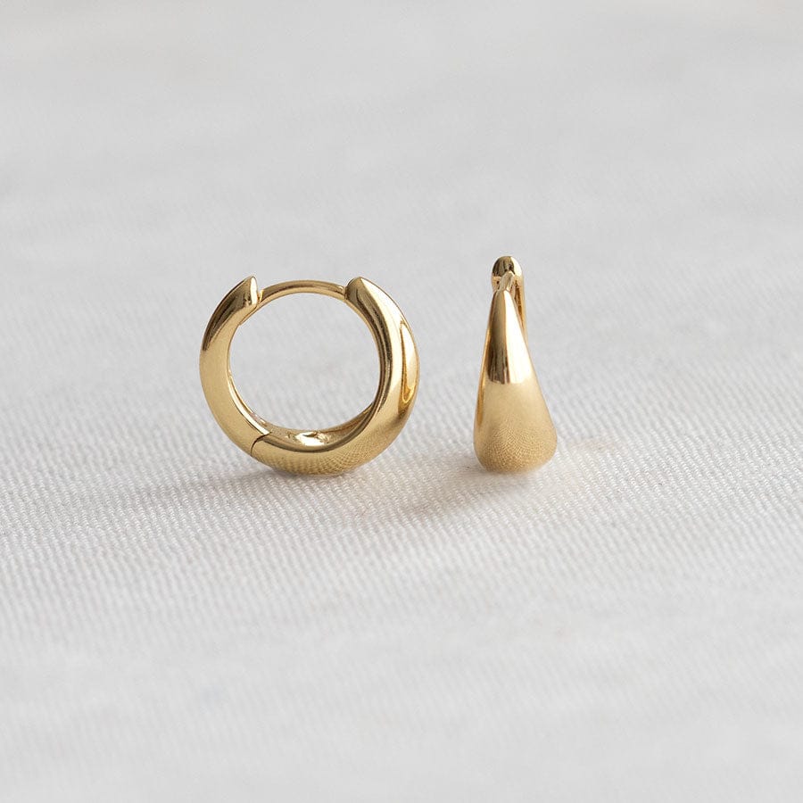A pair of gold-toned earrings on a light gray textured fabric background, featuring one small hoop earring and one teardrop-shaped stud earring.