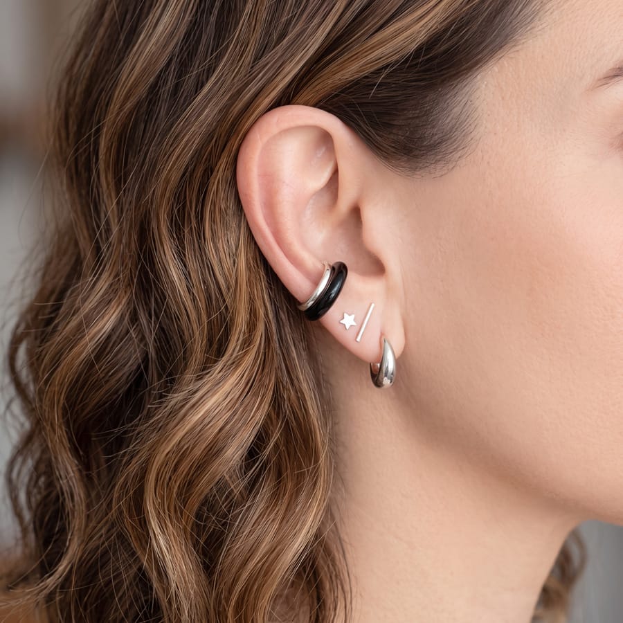 Close-up of a woman's ear wearing multiple small hoop and stud earrings, including a black hoop, a silver hoop, a silver star stud, and a silver diagonal stud.