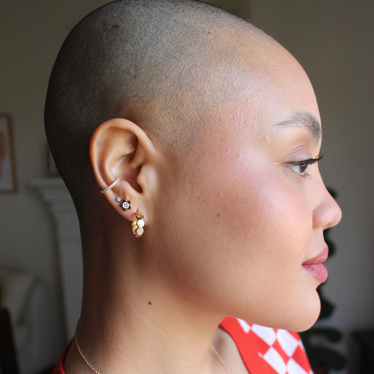 A model with a shaved head and beautiful smiley face wears the happy and sad hoop earrings on first piercing, and the smiley daisy enamel stud on second piercing with the CZ diamond stud in third piercing and a pave crystal ear cuff on top.