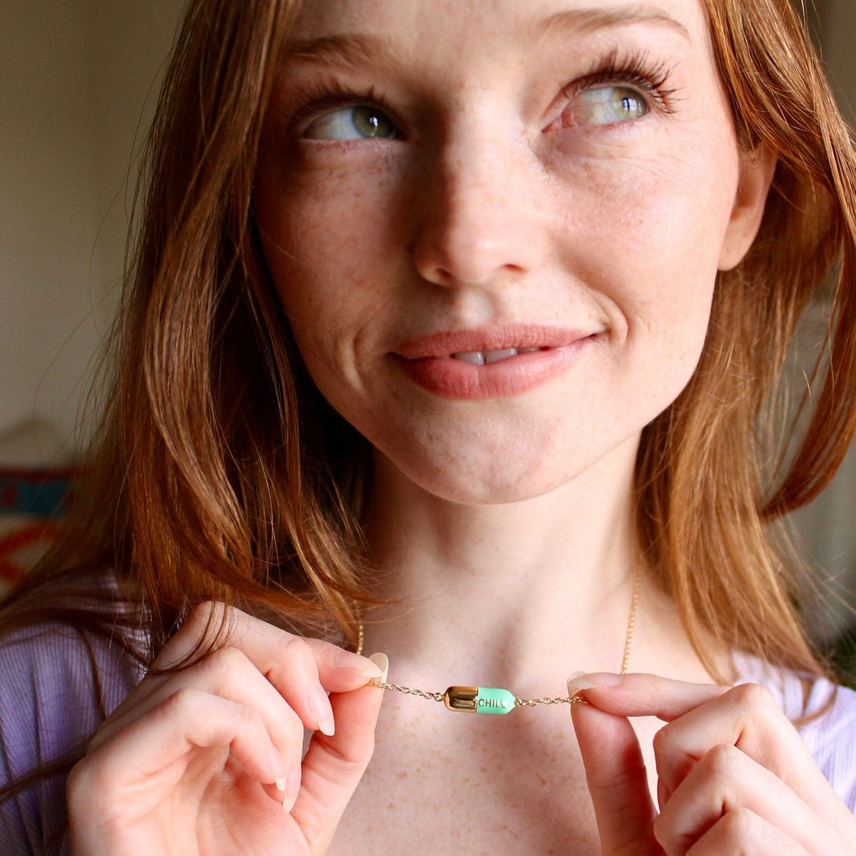Smiling woman with long red hair holding up the Big Chill Pill Chain Necklace