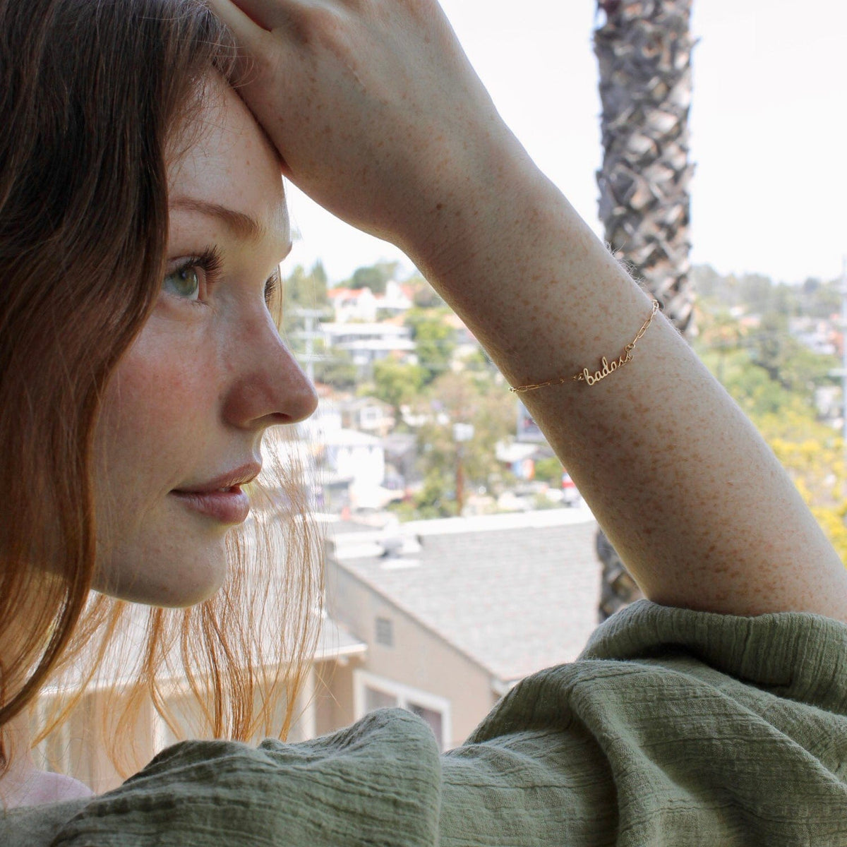 Woman with red hair resting her hand on her head, wearing the gold Badass Bracelet