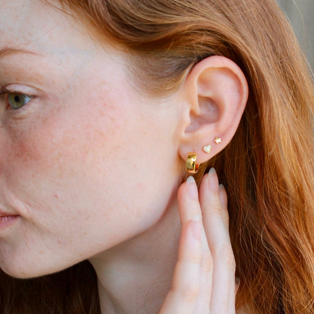 Woman with red hair, hand at her neck, wearing a gold Wide Hinged Huggie Hoop