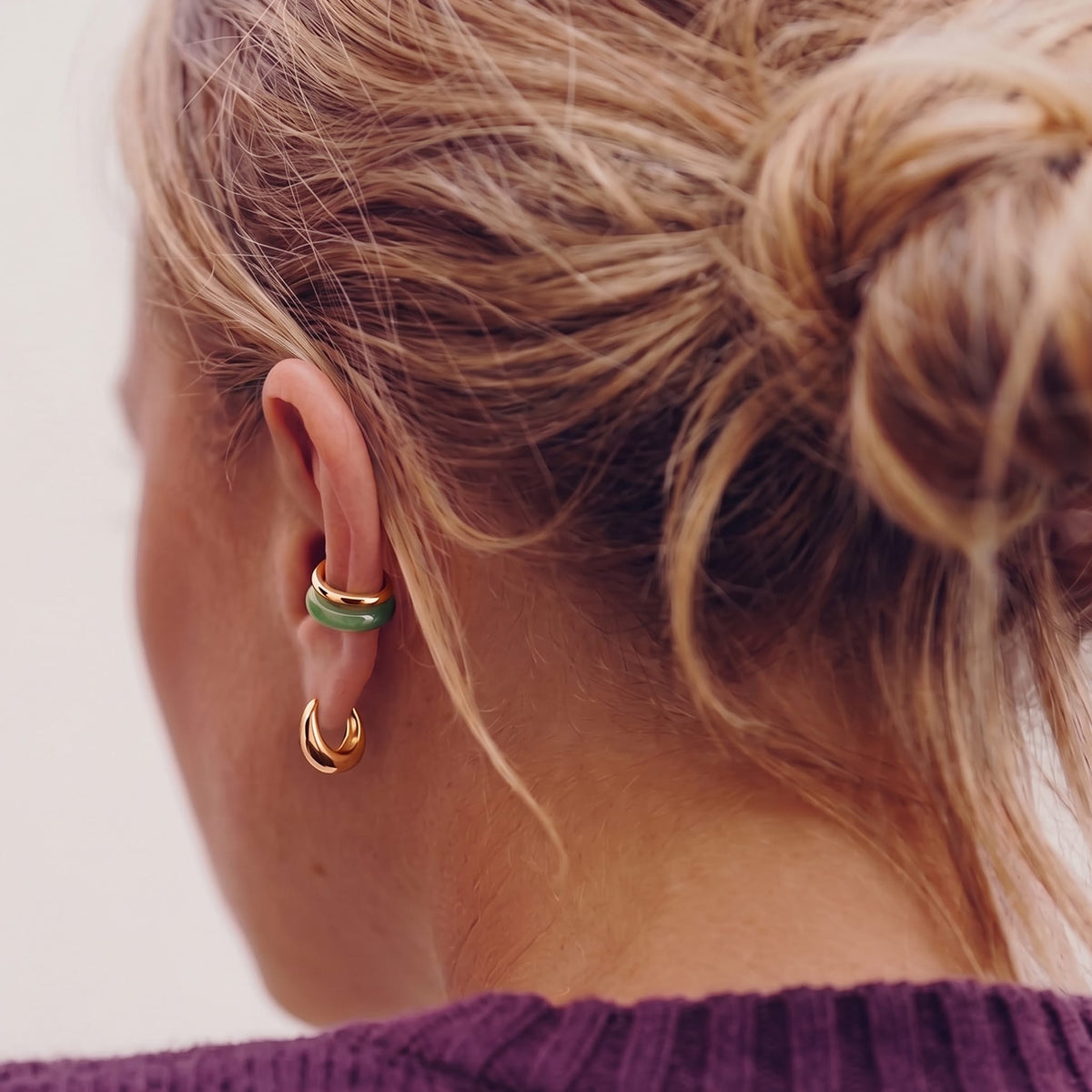 A close-up of a person's ear wearing two small gold hoop earrings, one with a thin green jade ear cuff and one with a gold ear cuff against a light neutral background.
