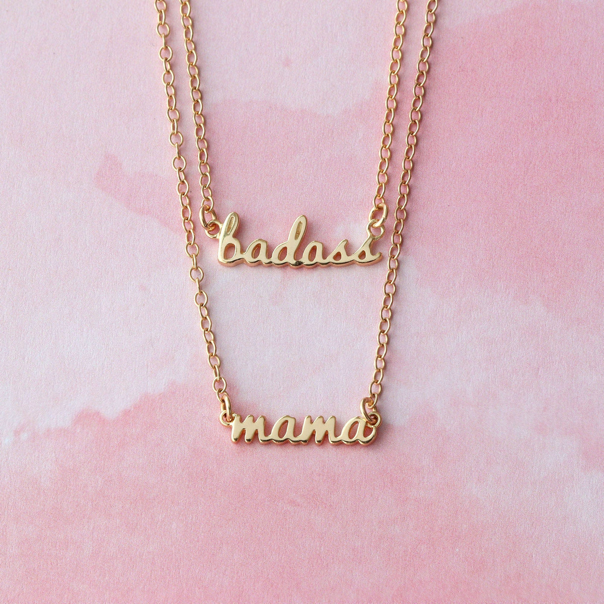 Gold Badass and Mama script charm necklaces layered on a pink background