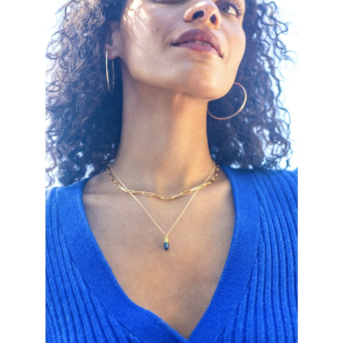 Woman with curly hair and gold hoops in a blue sweater wearing the Chill Pill Enamel Necklace layered with a gold chain