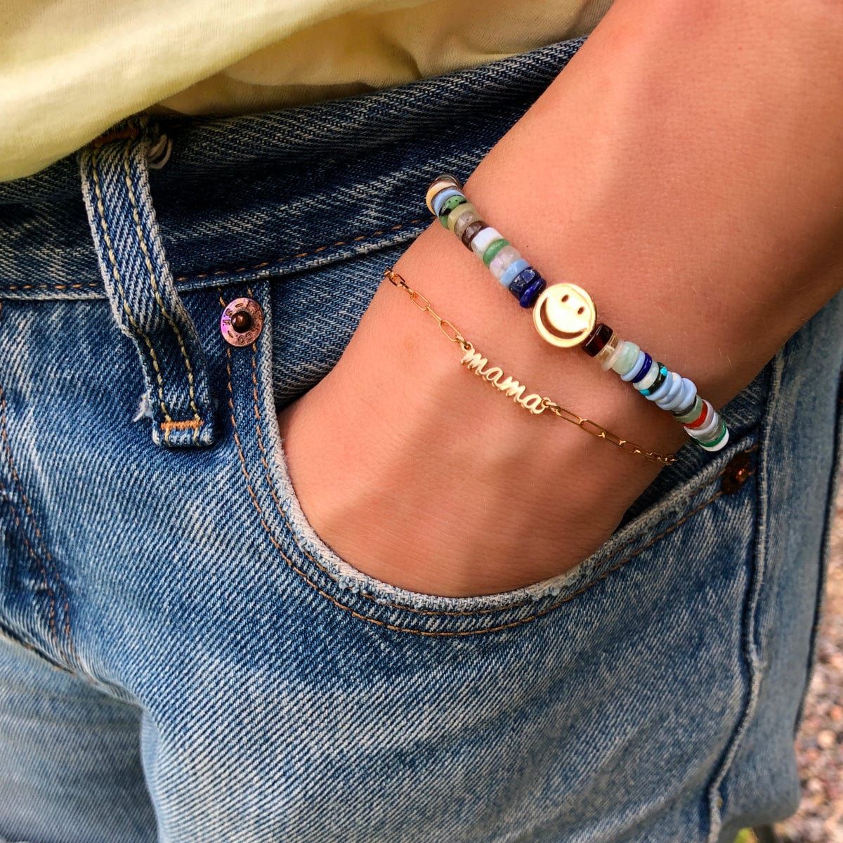 Hand in a jeans pocket wearing the gold Mama Bracelet with a beaded smiley face bracelet