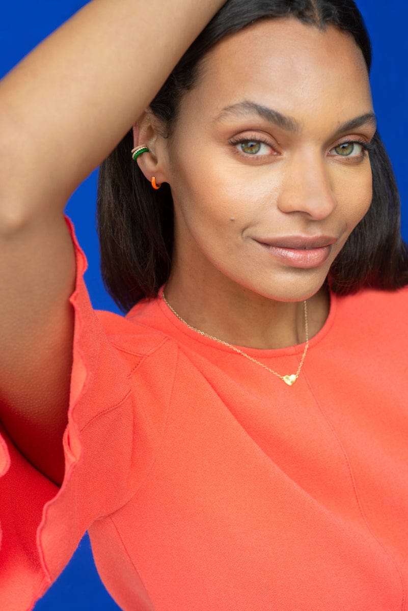 Smiling woman in an orange top wearing Enamel Huggie Hoops and a gold necklace
