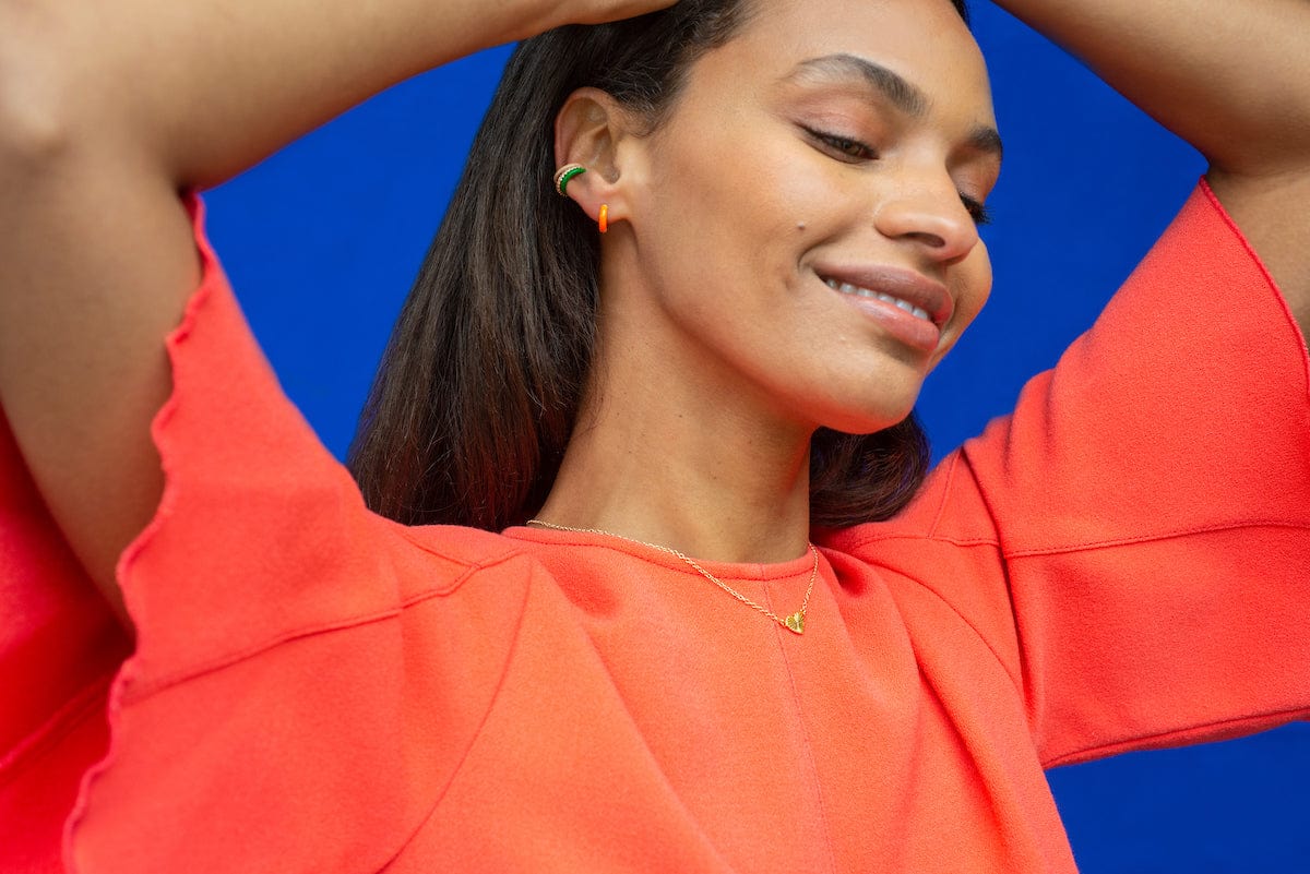 Smiling woman in an orange top, arms raised, wearing a Palette Enamel Ear Cuff