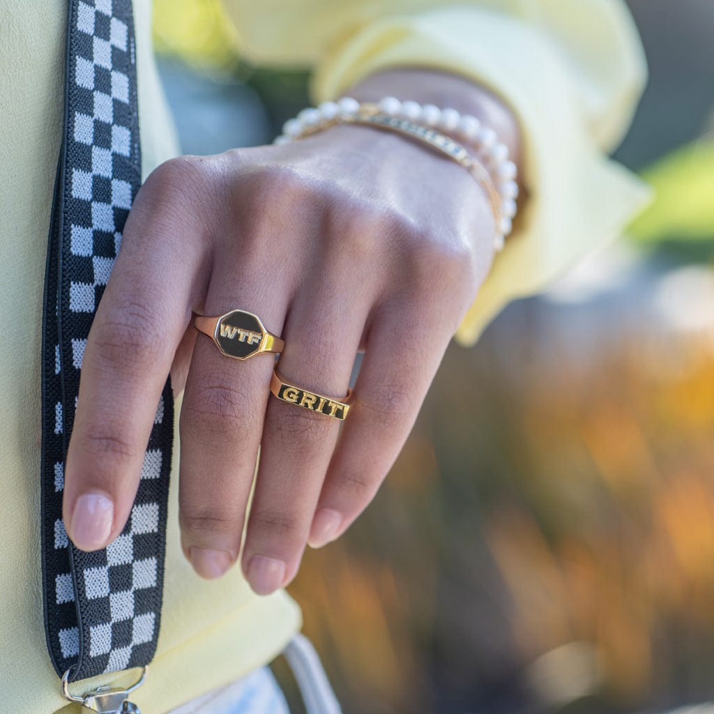Hand holding a checkered strap wearing the Grit Engraved Ring with other rings and pearl bracelets