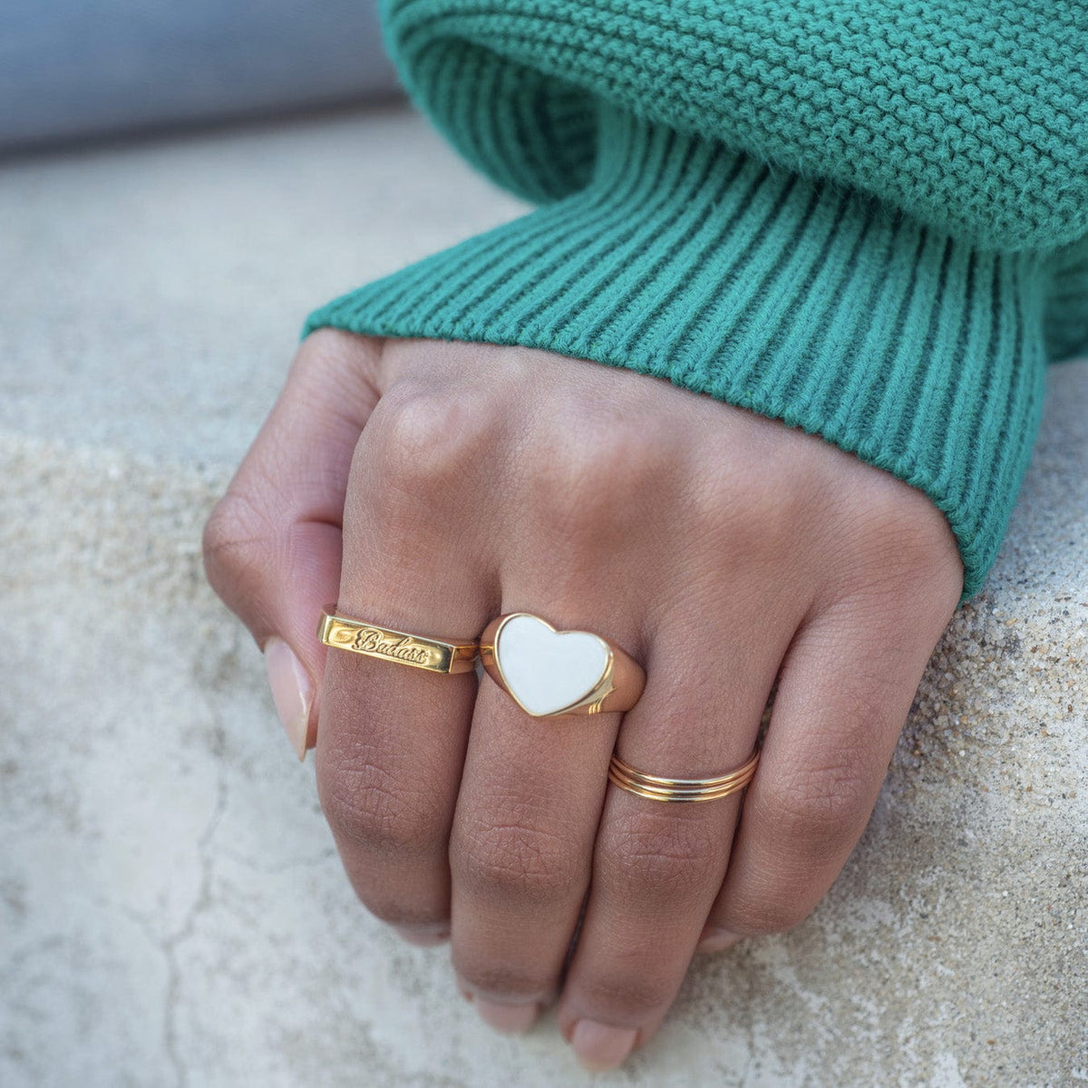 Hand in a green sweater wearing the Badass Script Engraved Ring with a white enamel heart signet